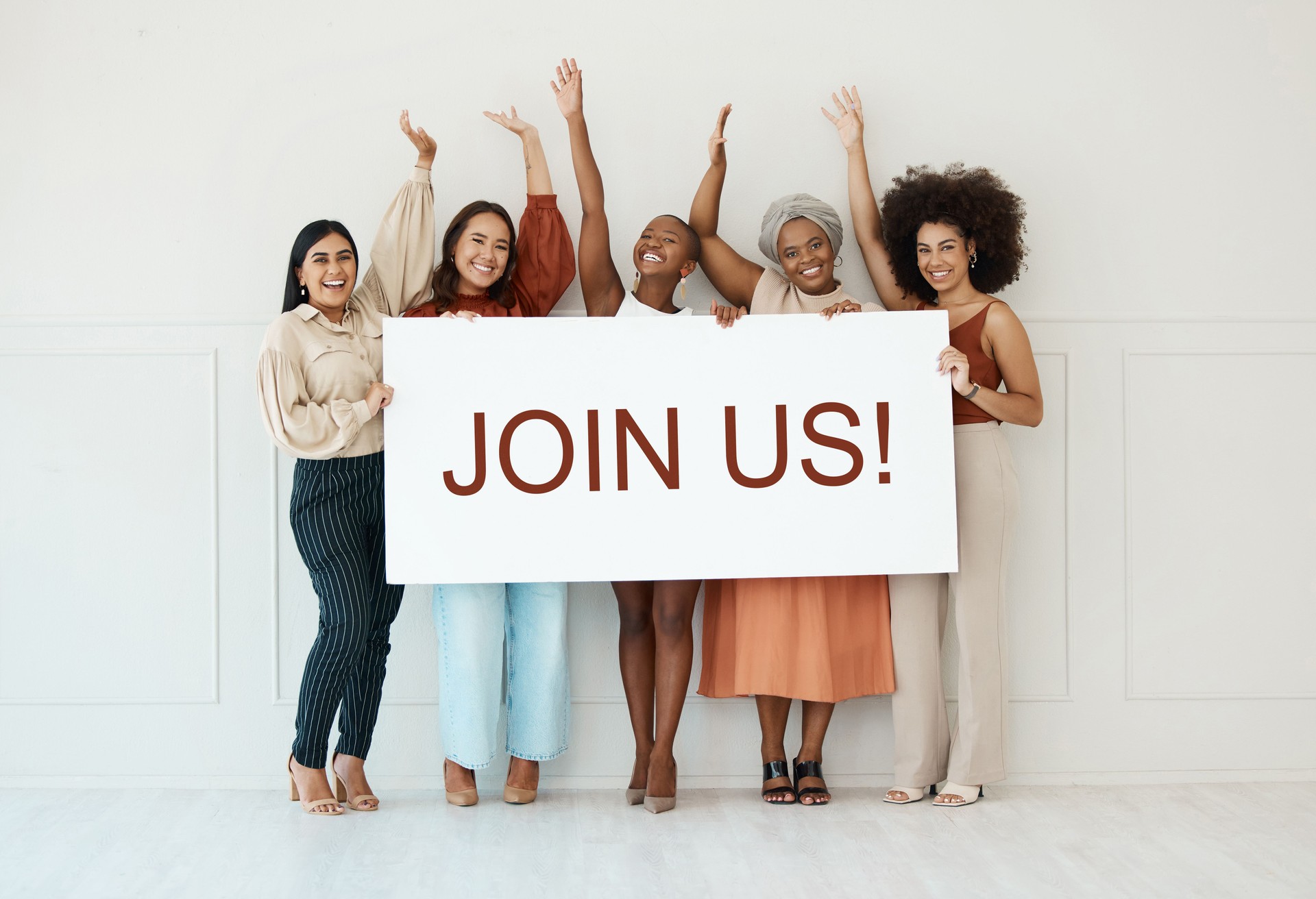 Group of five diverse young businesswomen standing against a wall in an office and holding a recruit sign. Happy colleagues holding a sign with a message while standing in a row together at work