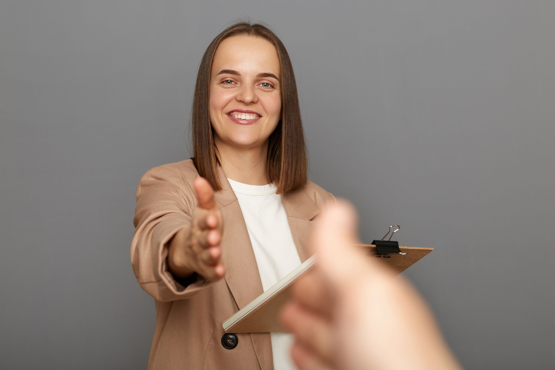 Portrait of attractive delighted positive brown-haired woman wearing jacket holding clipboard with documents, handshaking, deal, POV, standing isolated over gray background.